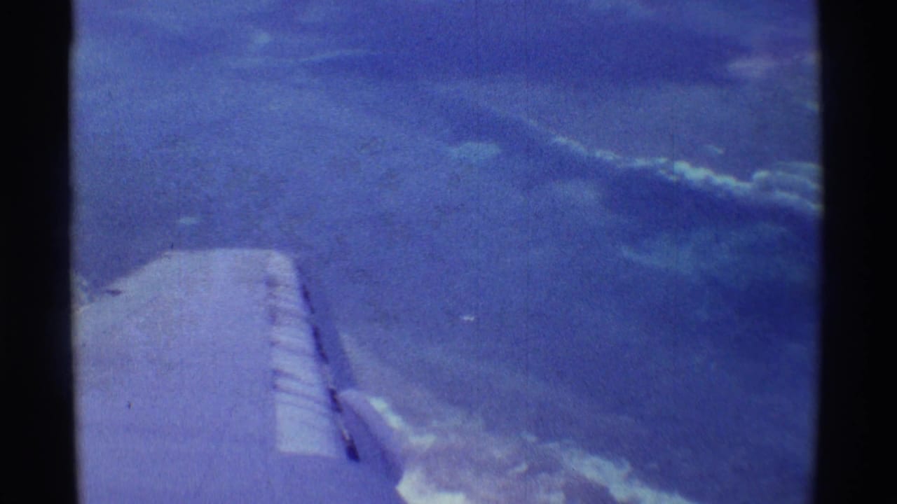 Home-movie view from a plane near Mauna Ulu, Hawaii, showing wing, ocean surface, cloud bands, and soft blue 8mm color.
