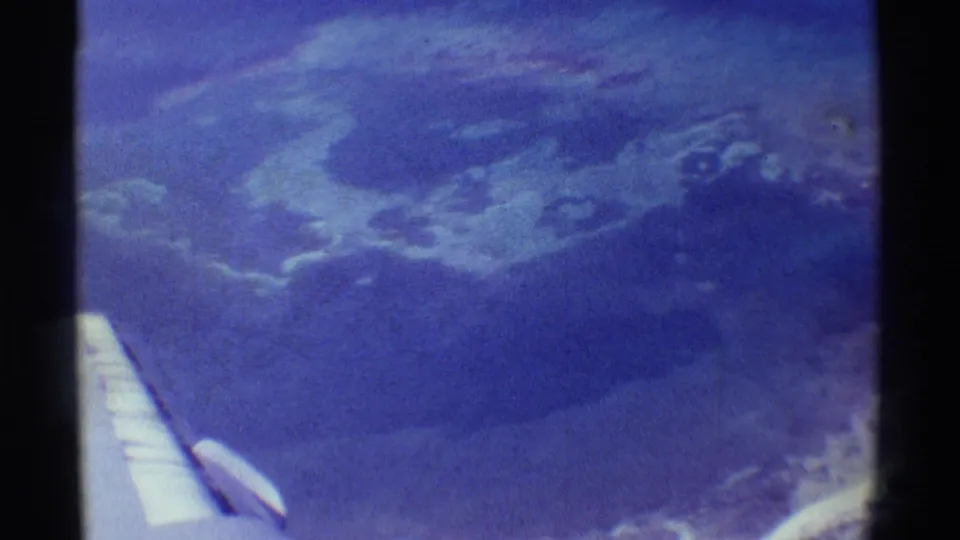 1971 8mm passenger view over Mauna Ulu, Hawaii, with an airplane wing at lower left and hazy volcanic terrain below.