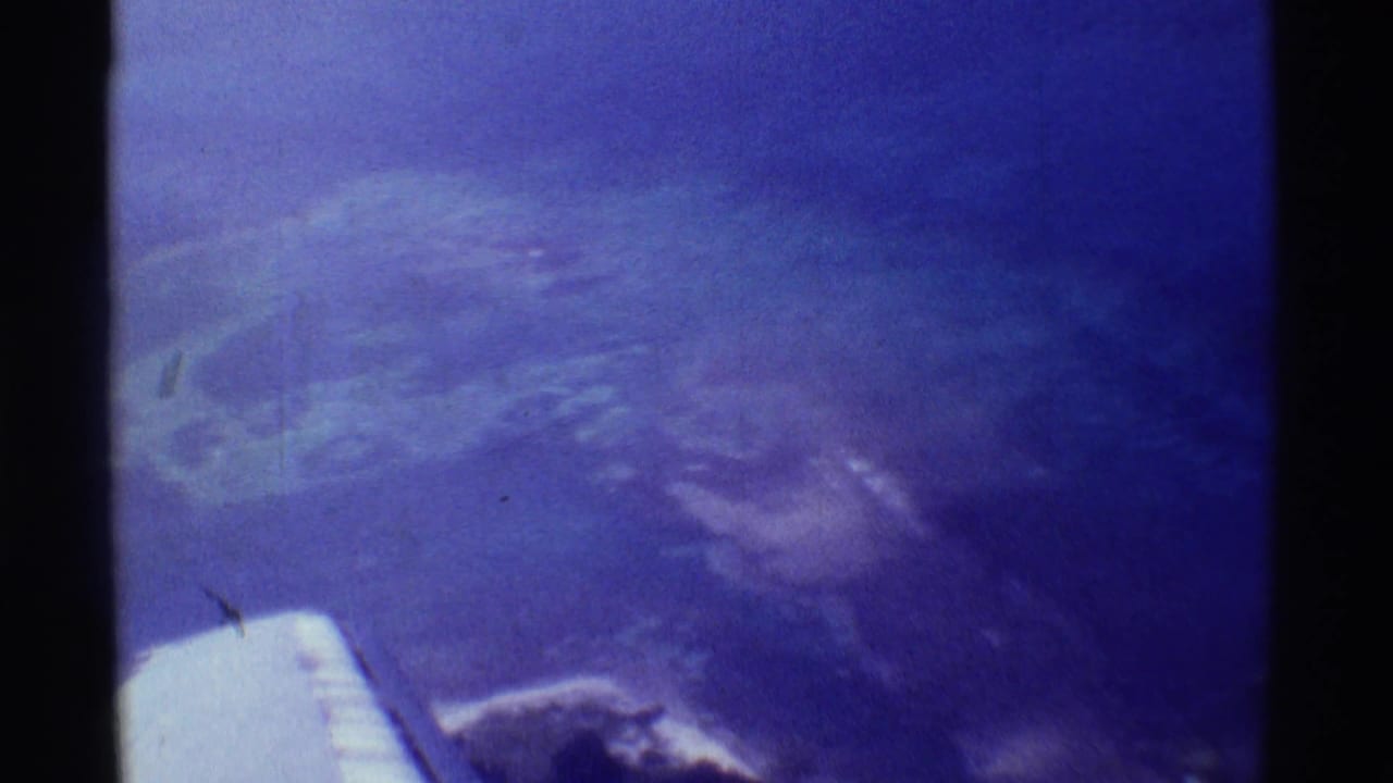 Soft 8mm frame from an airplane over Mauna Ulu, Hawaii, with hazy volcanic ground, cloud cover, and the wing tip near the bottom.