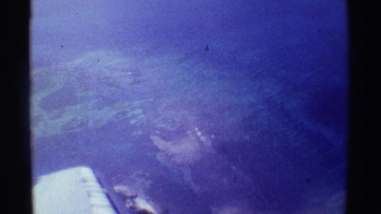 Airplane wing below a blue aerial view of ocean and land through aged film.