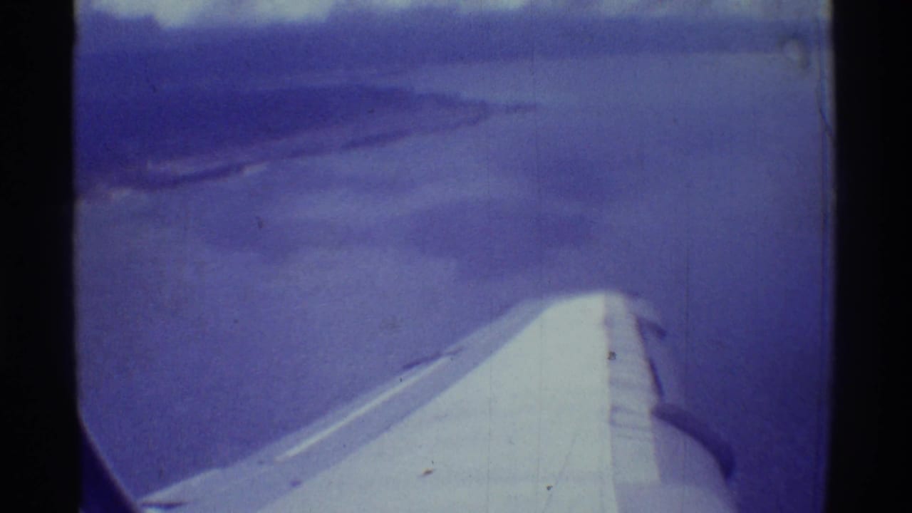 Aircraft wing over blue water with a distant shoreline or landform ahead.