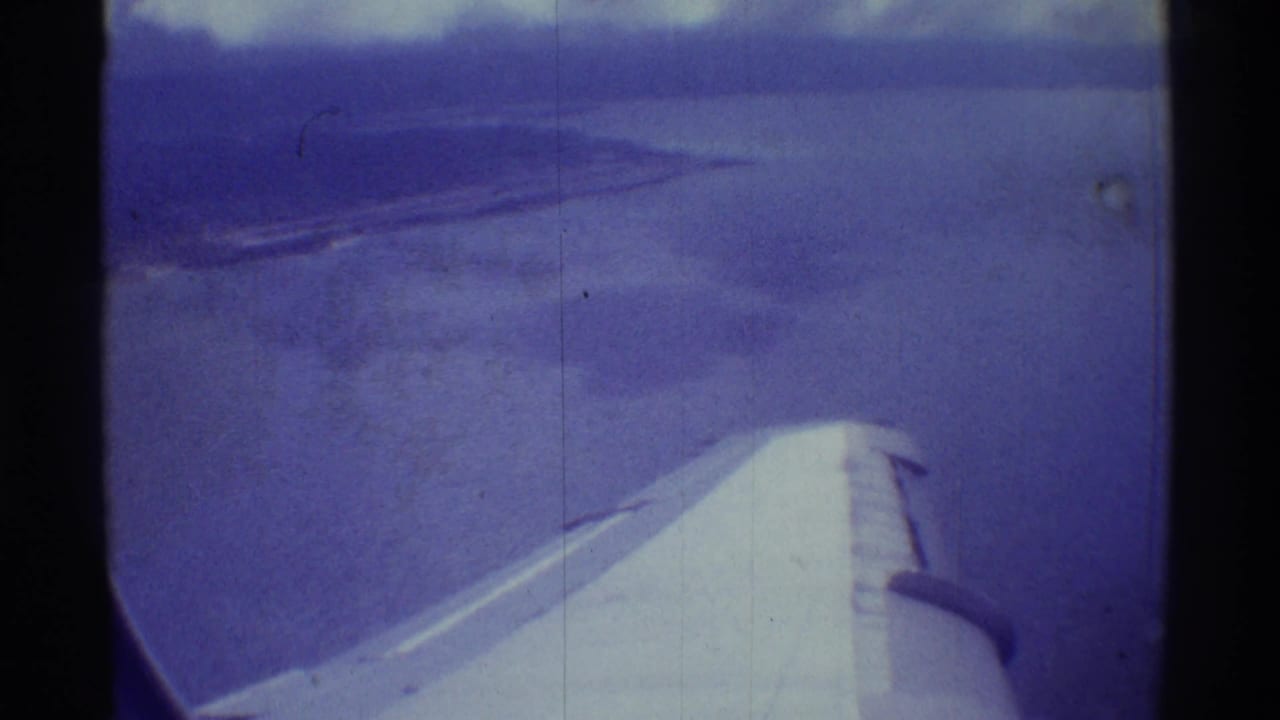 1971 8mm aerial view near Mauna Ulu, with a passenger-plane wing beneath blue ocean and a soft distant coastline.
