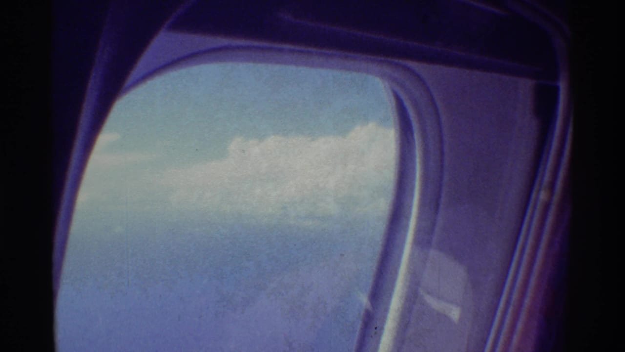 Inside a passenger aircraft, a rounded window frames clouds, blue sky, and part of the wing in 1971 Hawaii home-movie footage.