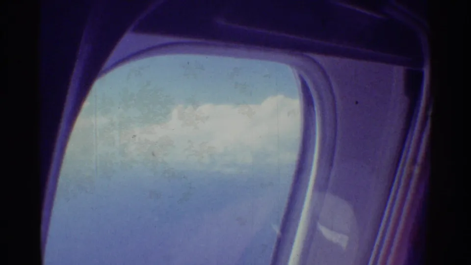 1971 8mm Hawaii travel frame showing a rounded airplane window with pale blue sky and soft clouds outside.