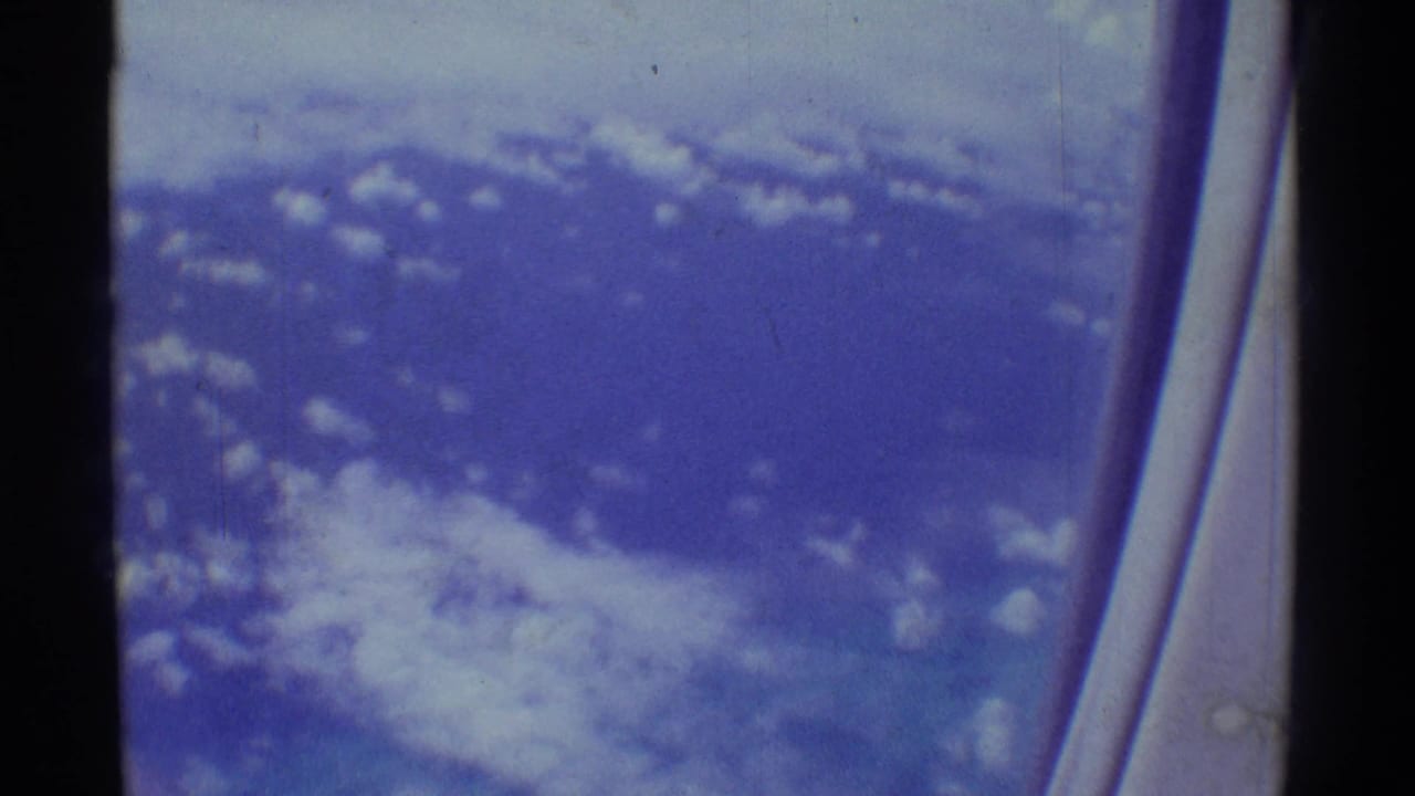 Aerial view over Hawaii from a passenger window, with scattered clouds and blue-toned terrain or sea below.