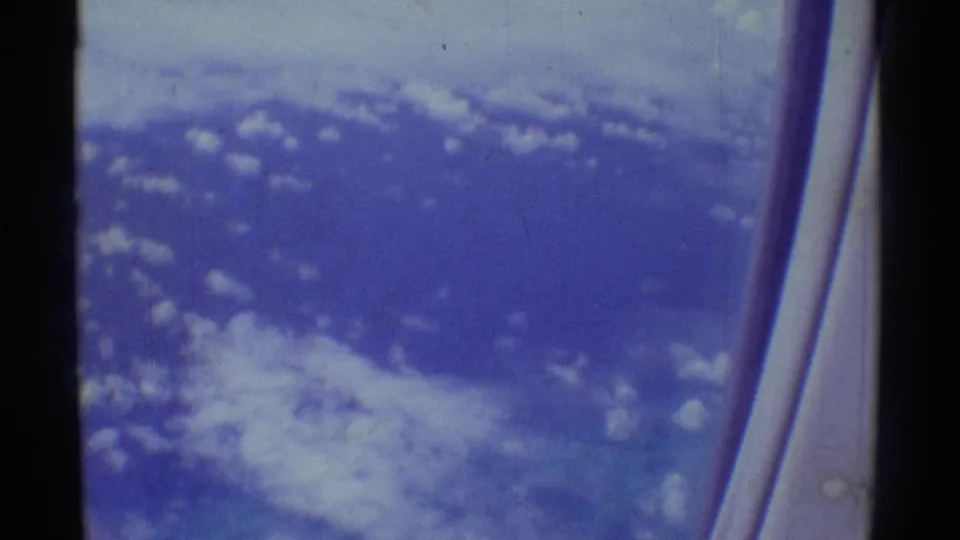 Aerial view over Hawaii from a passenger window, with scattered clouds and blue-toned terrain or sea below.