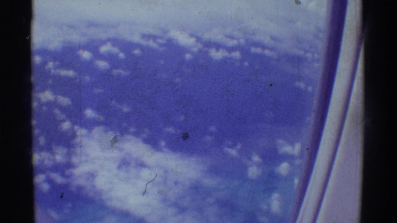 1971 8mm passenger view with scattered cloud cover and the curved edge of an airplane window at right.