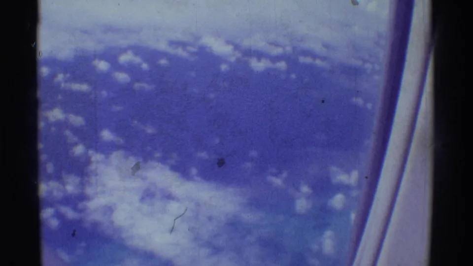 1971 8mm passenger view with scattered cloud cover and the curved edge of an airplane window at right.
