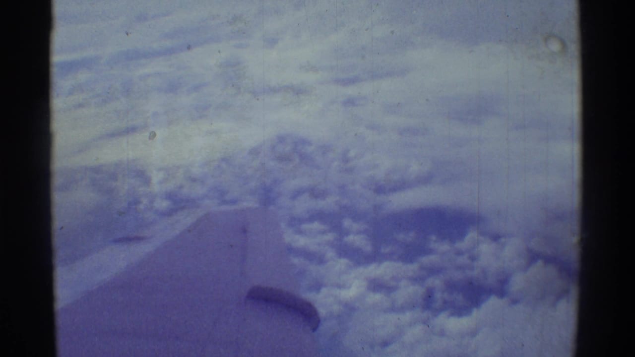 Passenger-window view over Hawaii with clouds below and a partial aircraft wing visible in soft 8mm color.