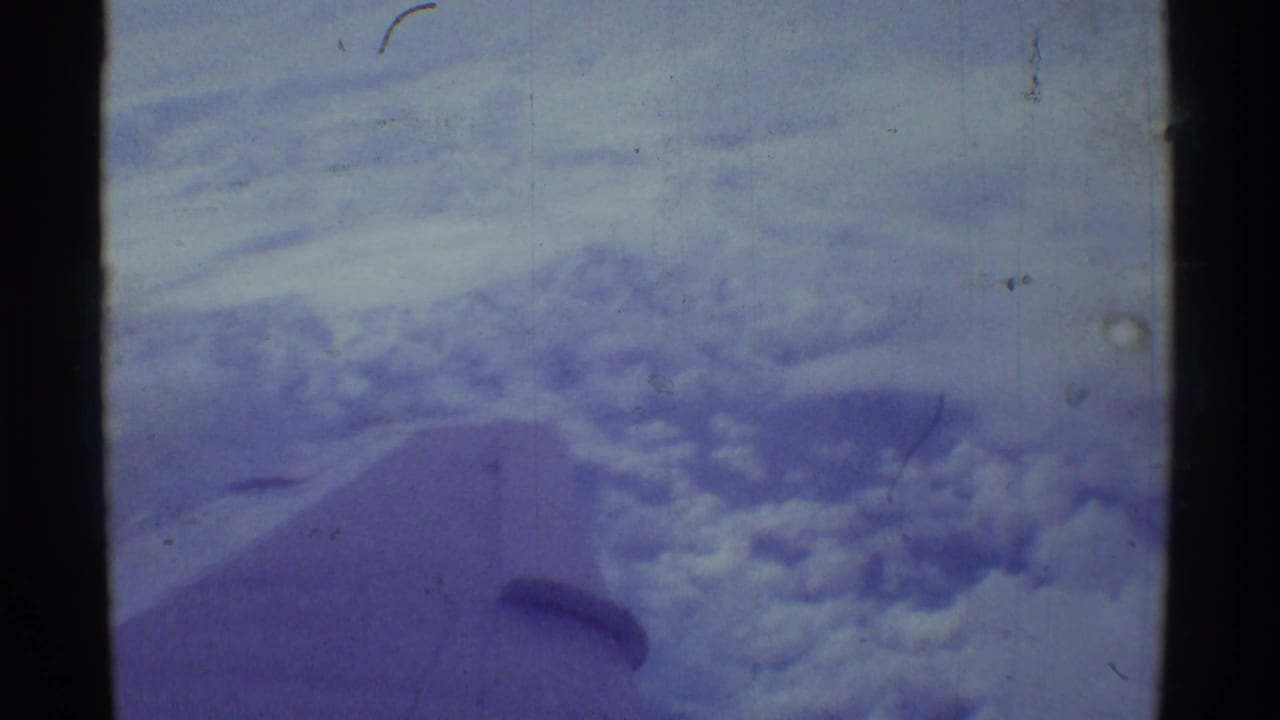 Aerial 8mm frame over Hawaii showing a cloud bank, blue sky, and the airplane wing edge within visible film borders.