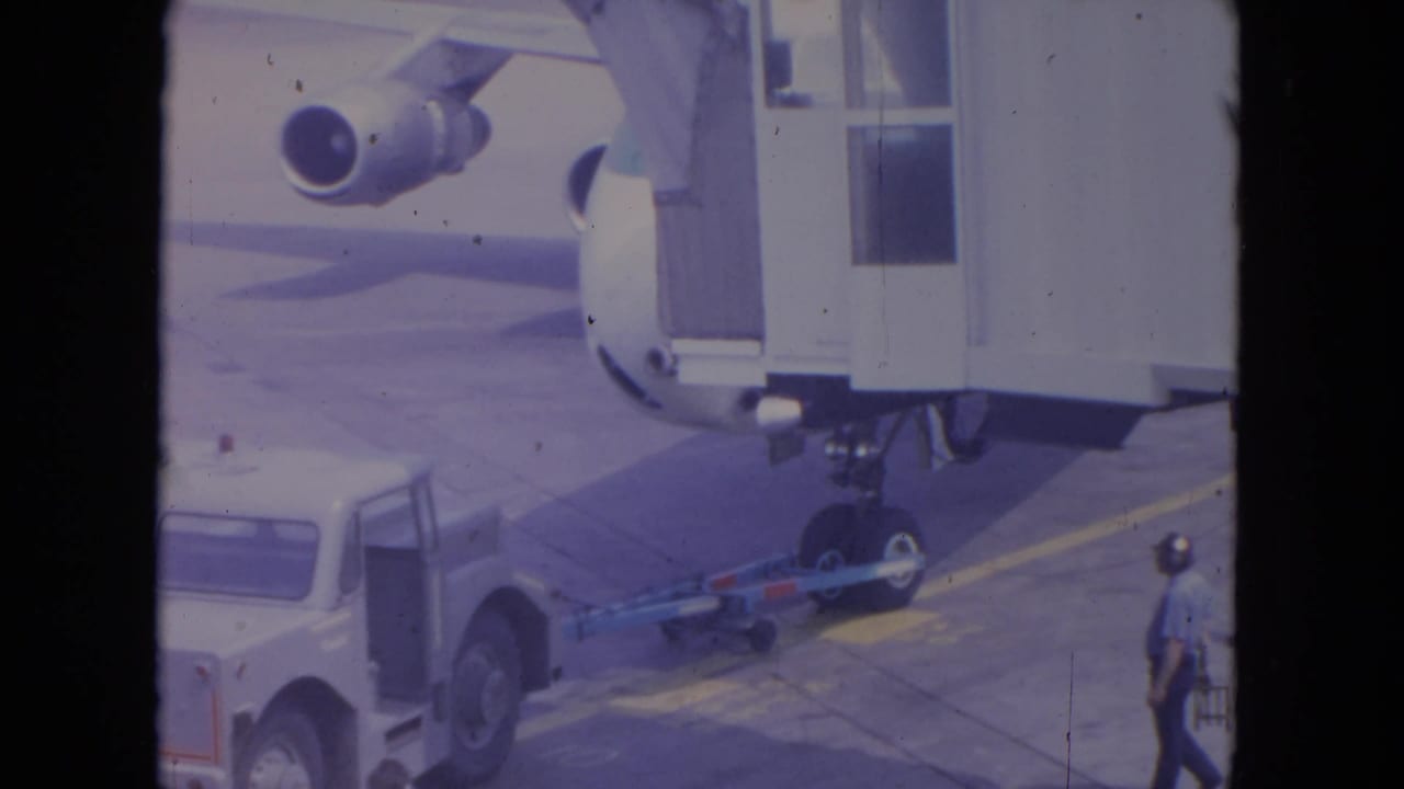 1972 O'Hare home-movie frame showing a jet bridge at a United aircraft door, with a tow vehicle and ground crew on the ramp.