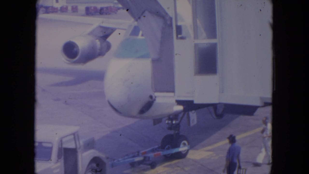 1972 O'Hare frame showing the United aircraft nose near a jet bridge, with tow equipment and a ground worker visible on the tarmac.