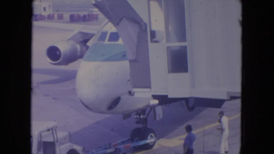 1972 O'Hare home-movie frame showing a blue-striped commercial jet nose beside a jet bridge with ground crew below.