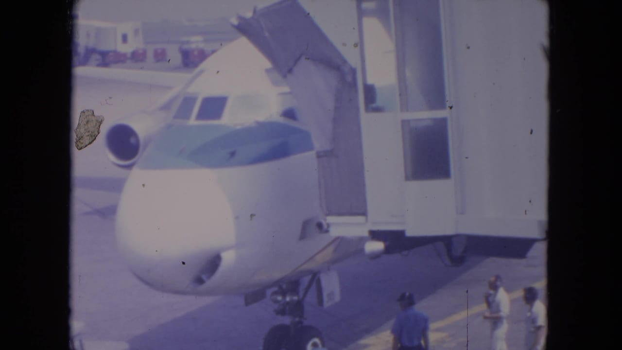 Home-movie view at O'Hare in 1972 showing the front of a blue-striped jet aligned with a gate jet bridge.