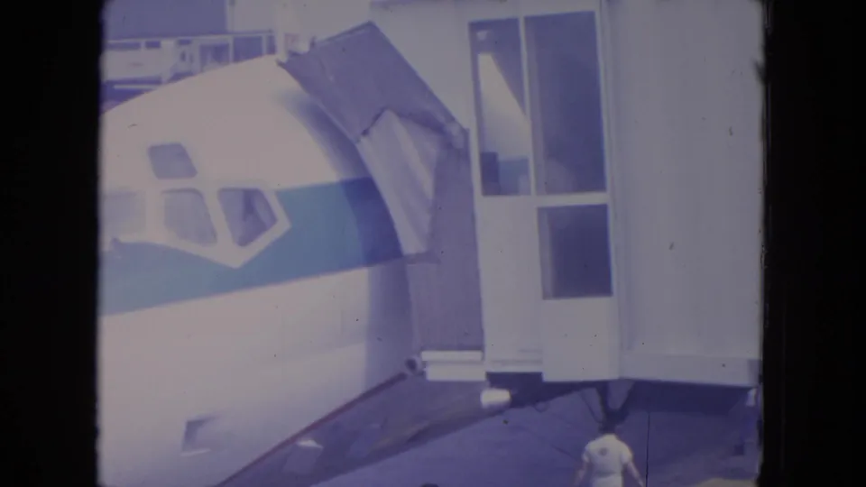 1972 O'Hare home-movie frame showing a white airliner with a blue stripe parked at a gate as the jet bridge stands near the front door.