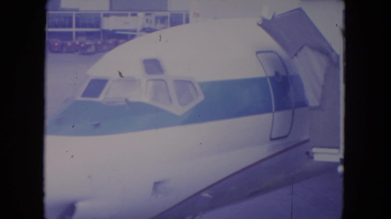 1972 O'Hare frame showing the front of a white airliner with blue stripe after the jet bridge has retracted from the doorway.