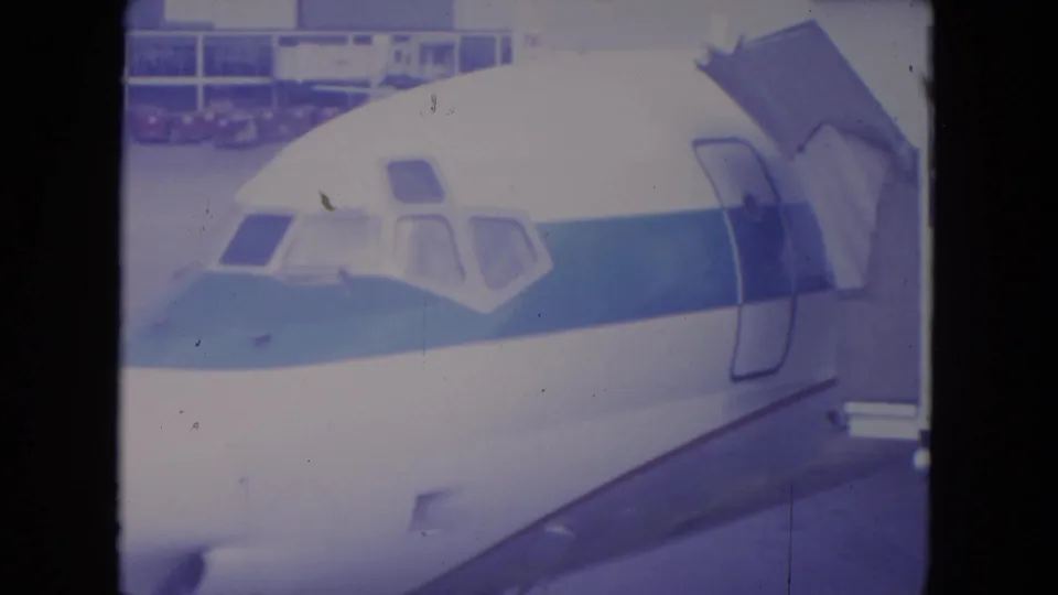 1972 O'Hare frame showing the front of a white airliner with blue stripe after the jet bridge has retracted from the doorway.