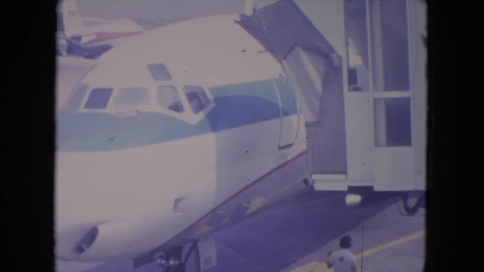 1972 O'Hare home-movie frame showing the nose of a commercial jet at a gate with a connected jetway and faint ground activity.