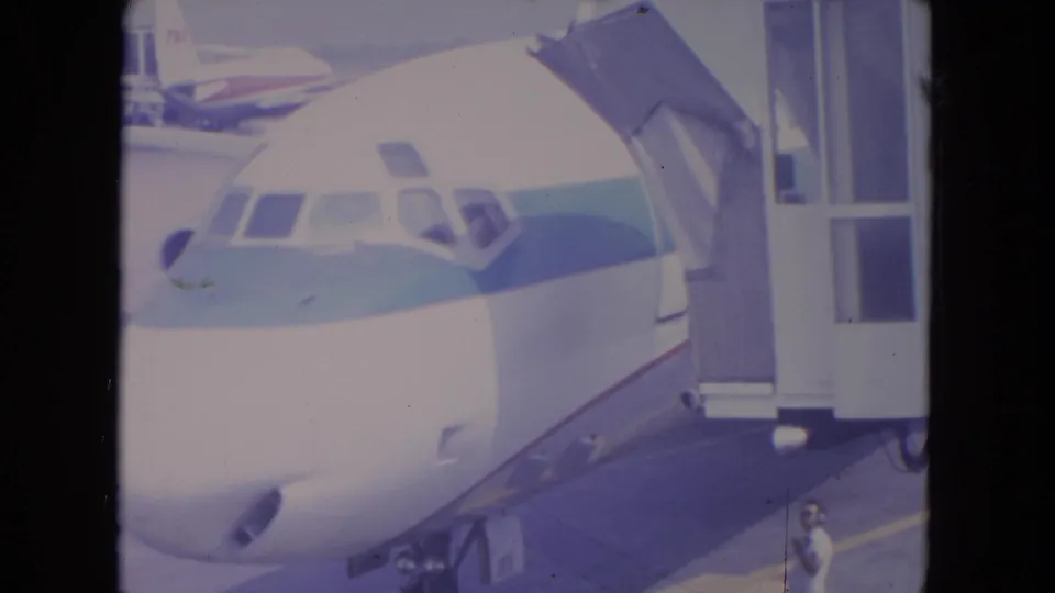 1972 O'Hare frame of a white commercial jet parked at a gate, with jetway equipment and a small visible person on the tarmac.