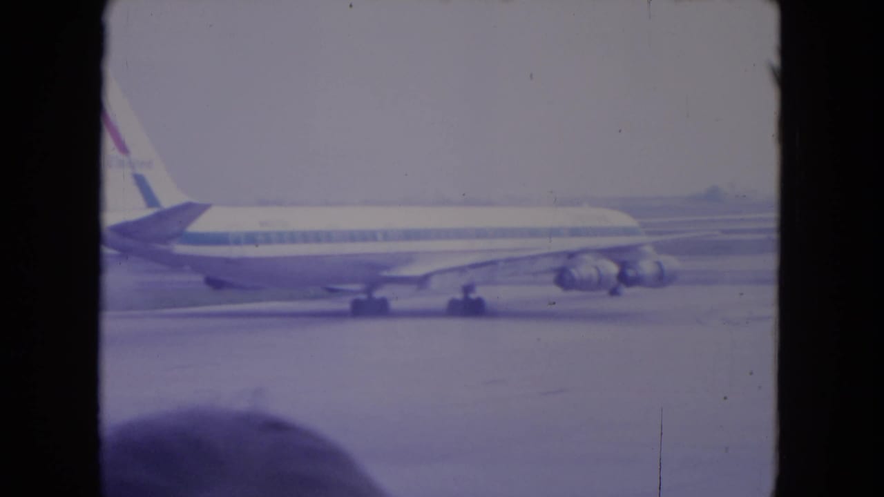 1972 O'Hare home-movie frame showing a commercial airliner taxiing in profile across the tarmac, softened by faded 8mm color.