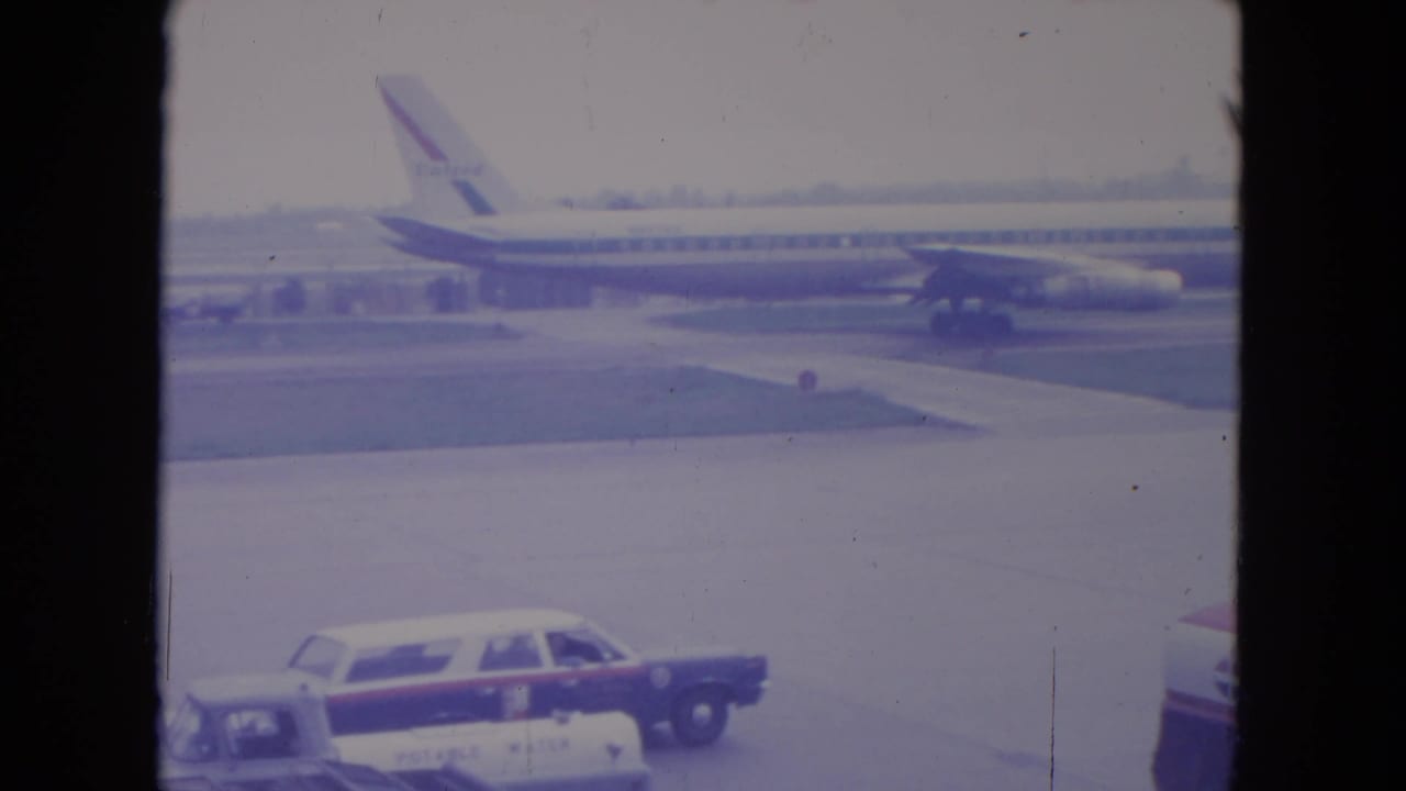 A United Airlines jet is seen across the O'Hare tarmac, with airport cars in the foreground and 8mm film scratches visible.