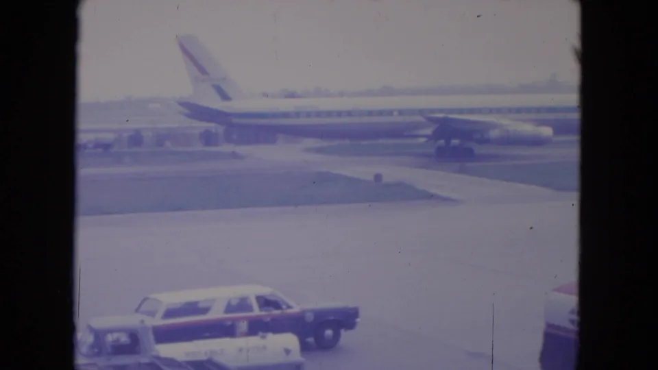 A United Airlines jet is seen across the O'Hare tarmac, with airport cars in the foreground and 8mm film scratches visible.