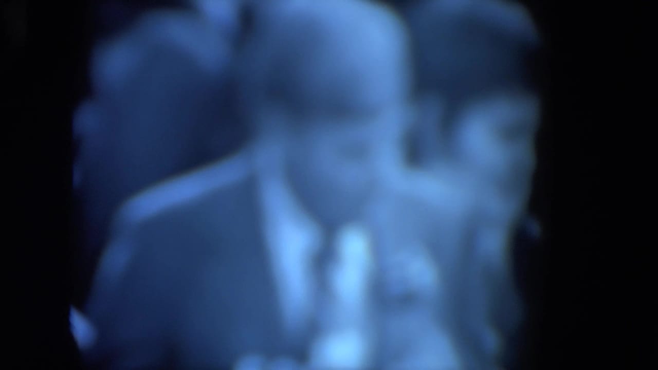 Blurry 1972 O'Hare Chicago 8mm frame showing a suited man seated near a desk-like surface, with blue cast and flicker.
