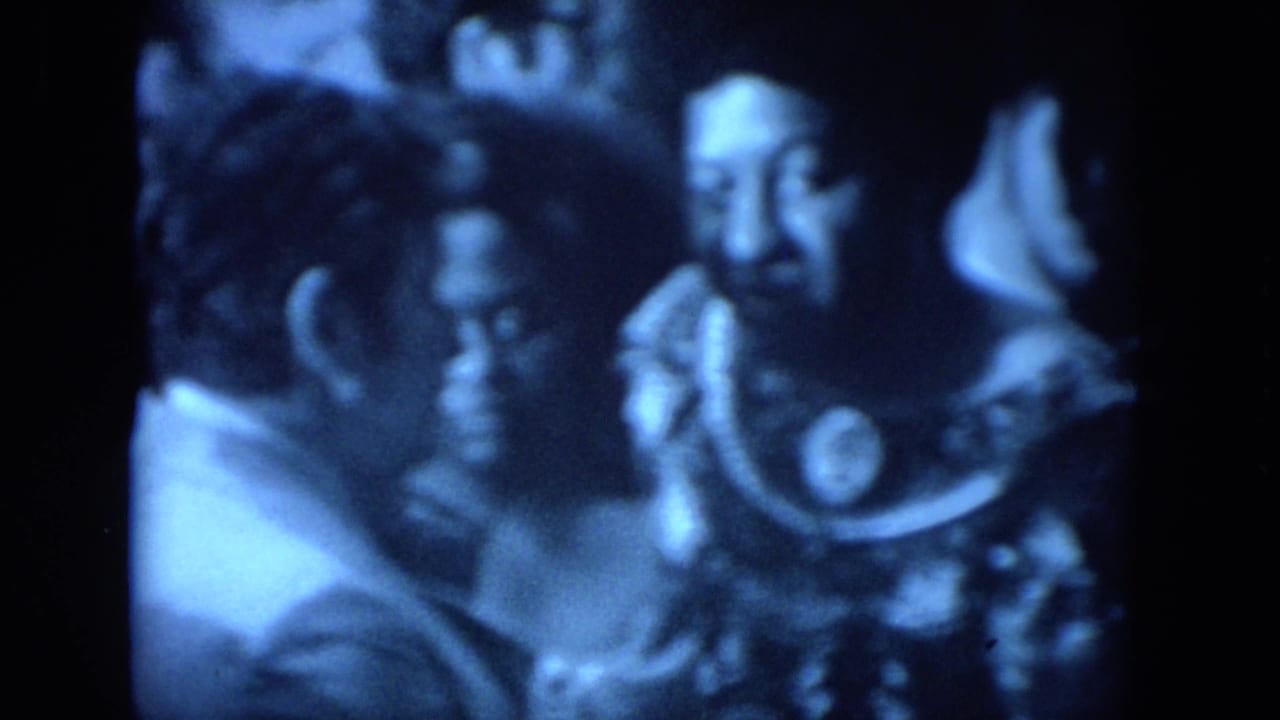 1972 O'Hare Chicago frame showing adults clustered around a bearded man who appears to be speaking near a microphone.