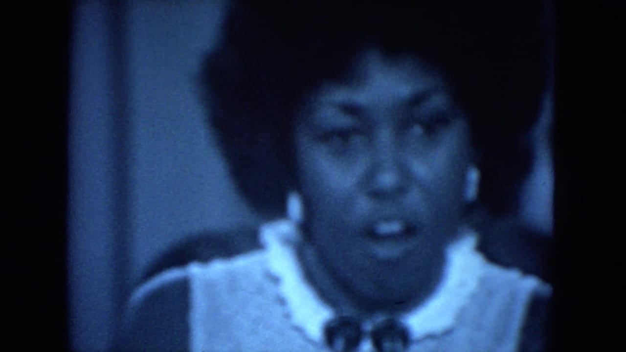 Close-up of a Black woman with an afro hairstyle speaking at the 1972 O'Hare Chicago event, rendered in soft blue 8mm film tones.