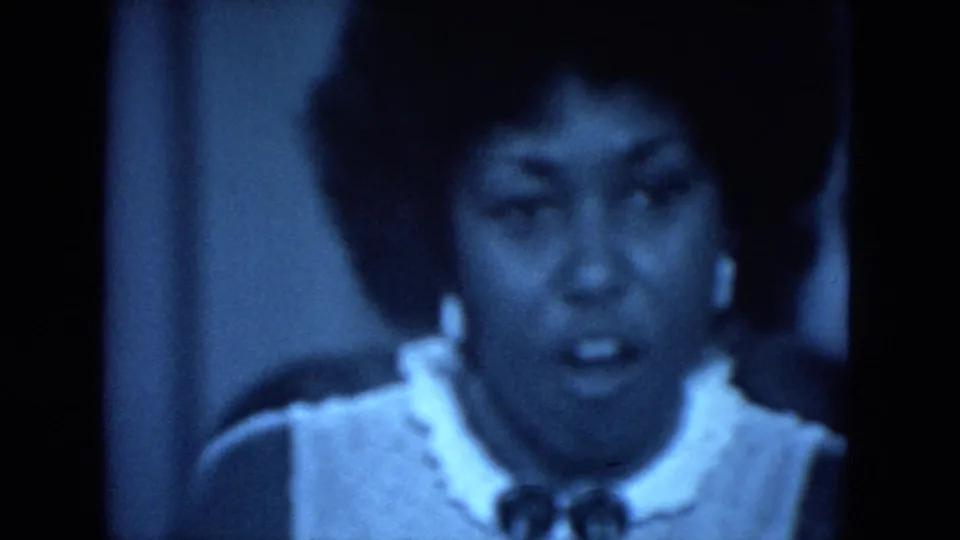 Close-up of a Black woman with an afro hairstyle speaking at the 1972 O'Hare Chicago event, rendered in soft blue 8mm film tones.