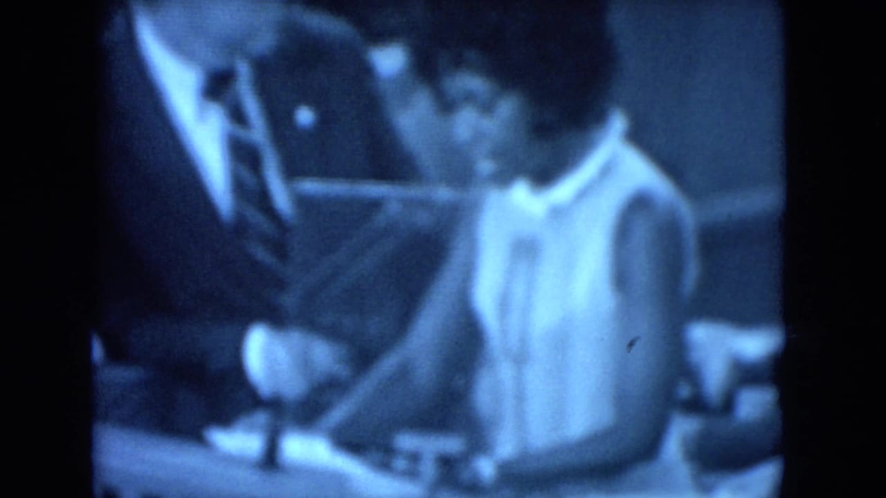 Blue-tinted 8mm frame showing the woman near a suited man and a table or lectern area during the 1972 O'Hare Chicago gathering.