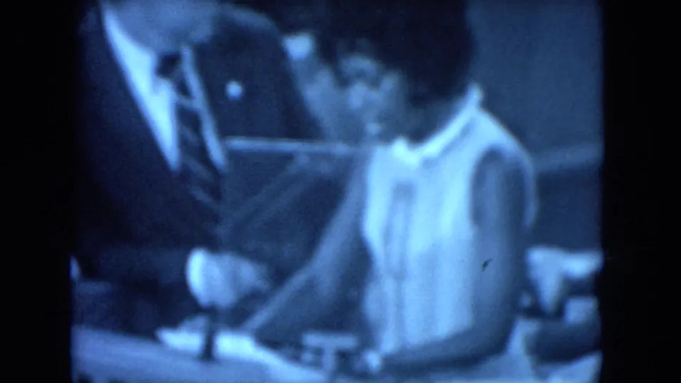 Blue-tinted 8mm frame showing the woman near a suited man and a table or lectern area during the 1972 O'Hare Chicago gathering.