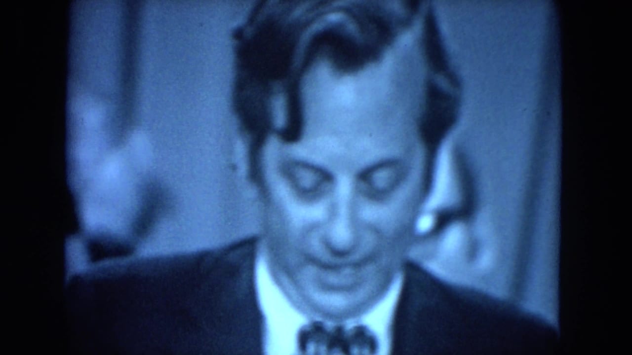Close-up of a suited man looking down while speaking or reading.