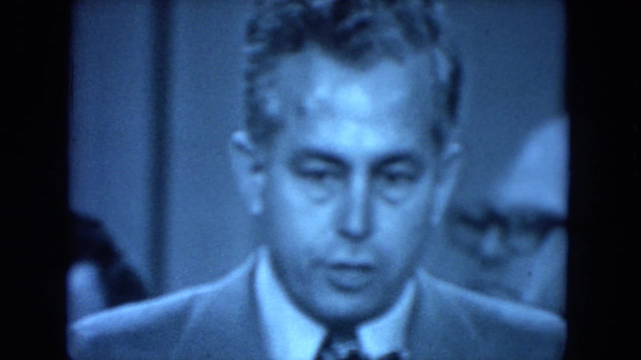 Blue-toned 8mm close-up of a suited adult man at the formal gathering, with another partially visible face behind him.