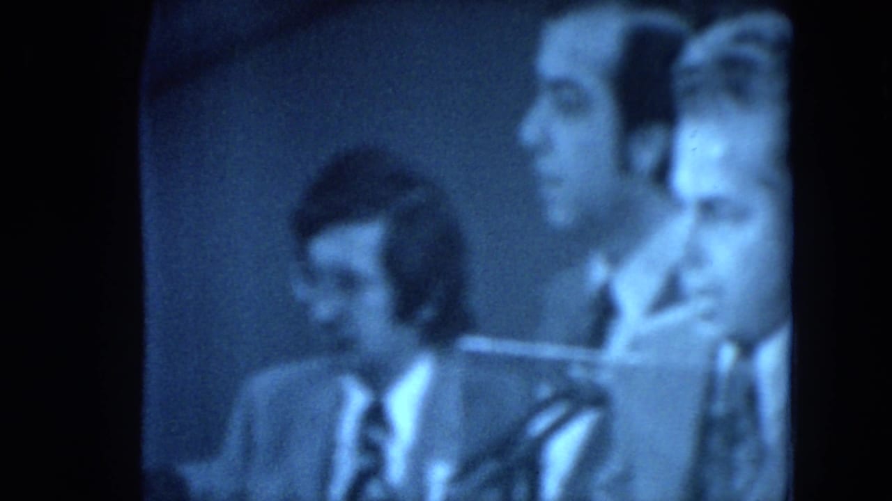Side-angle 1972 8mm frame showing suited men near the front of a formal gathering, with faces partly blurred by motion and focus.