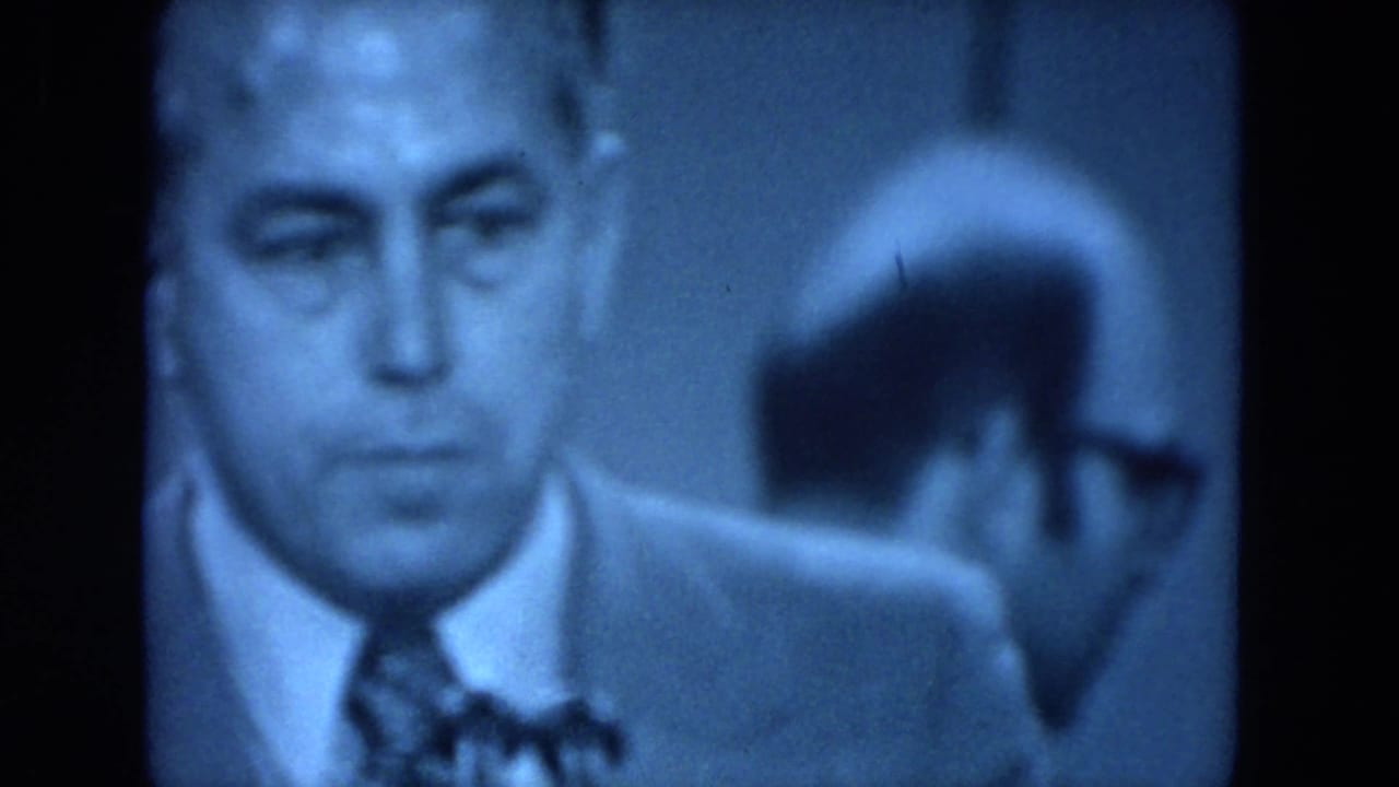 Blue-tinted 1972 O'Hare Chicago 8mm frame showing a suited man in close view during a formal indoor gathering.