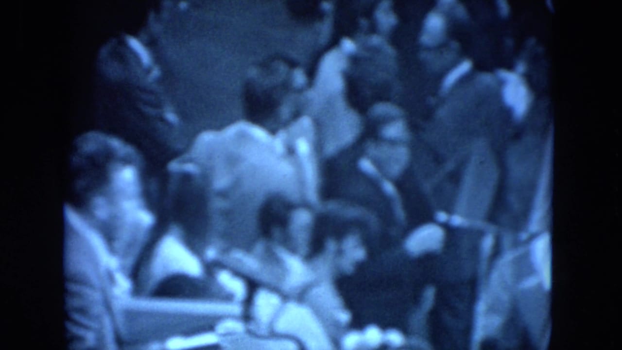 Blue-tinted 1972 meeting footage showing a crowded indoor group, likely listening or waiting during proceedings.