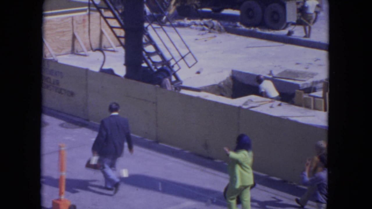 Pedestrians walk beside a construction barrier as a woman in green enters the frame.