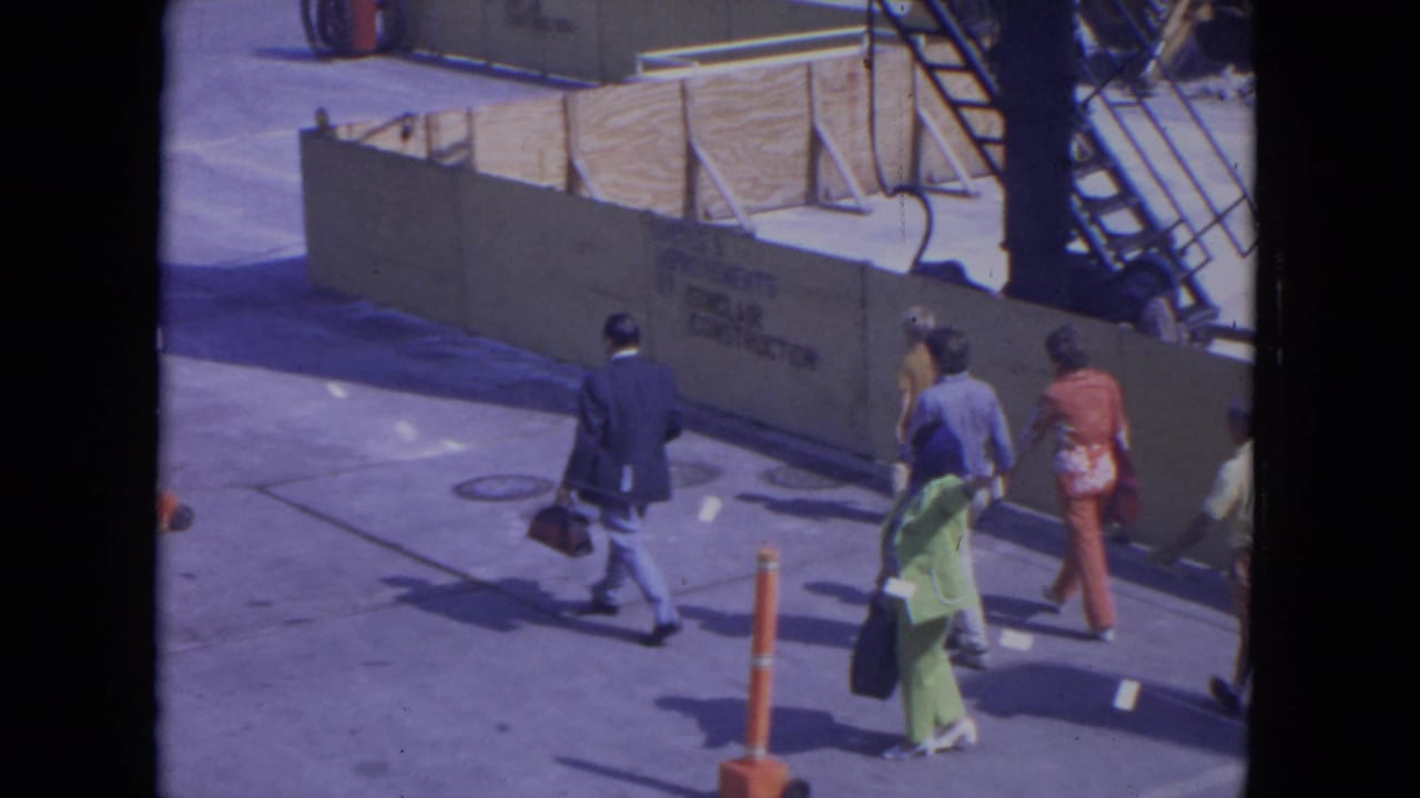 Travelers in bright clothing walk across an airport tarmac beside a construction barrier.