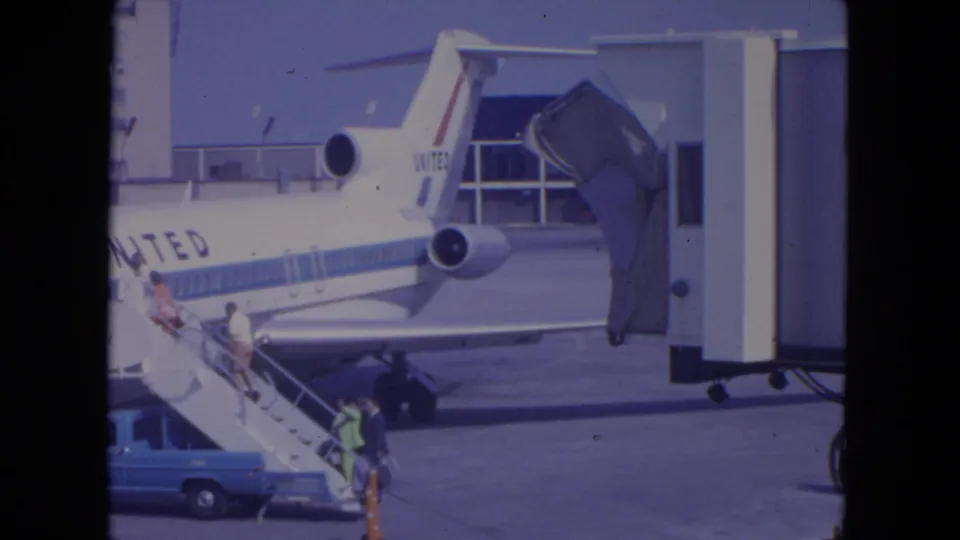 1972 O'Hare home-movie frame showing passengers boarding a United Airlines jet by mobile stairs beside a jet bridge.