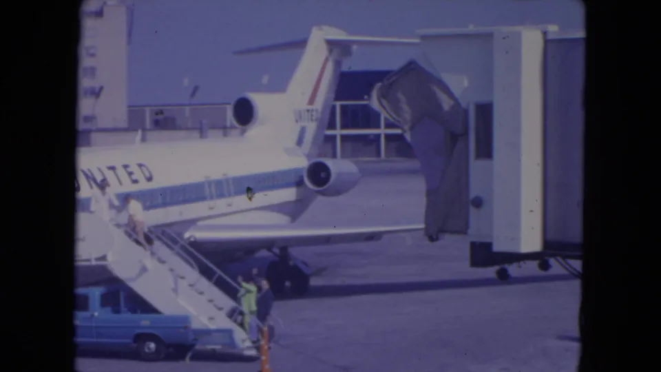 1972 O'Hare frame with a United Airlines jet, mobile stairs, passengers boarding, a blue service truck, and terminal gate equipment.