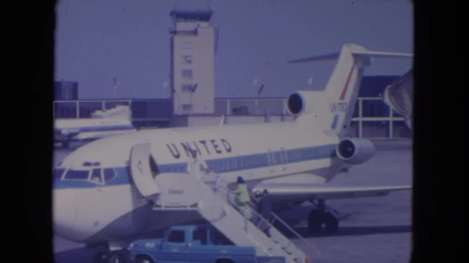 1972 home-movie view at Chicago O'Hare showing a United Airlines jet with boarding stairs and people on the tarmac.