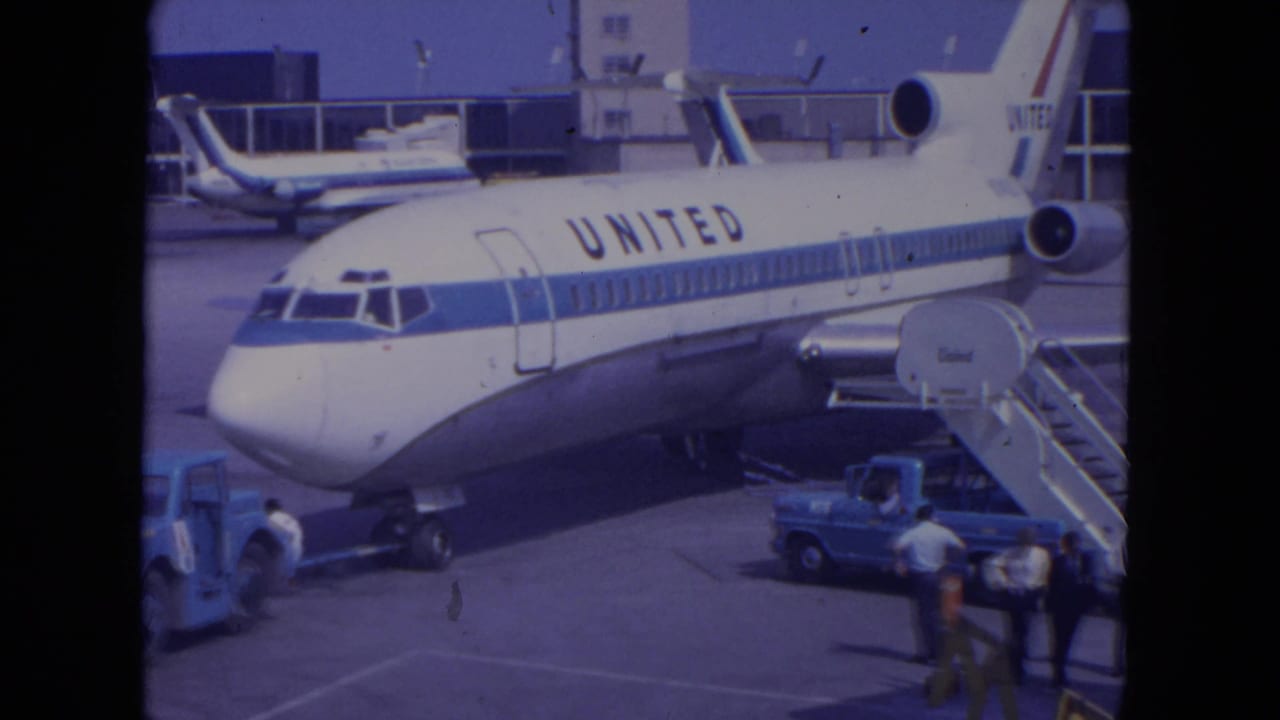 1972 O'Hare home-movie frame showing the United jet's front section, boarding stairs, ramp vehicles, and people standing below.