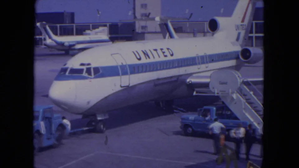 1972 O'Hare home-movie frame showing the United jet's front section, boarding stairs, ramp vehicles, and people standing below.