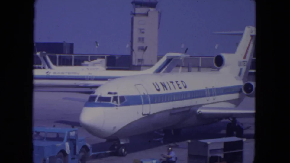 1972 O'Hare home-movie frame showing a United jet on the tarmac, airport tower behind it, and an Eastern aircraft in the background.