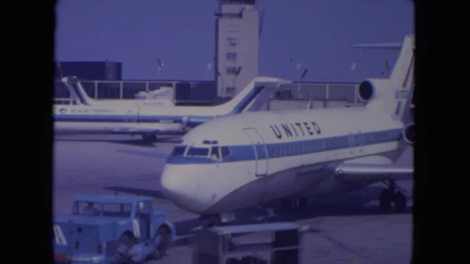 United aircraft nose and fuselage at O'Hare in 1972, with airport service vehicles, tower structures, and another plane visible behind.
