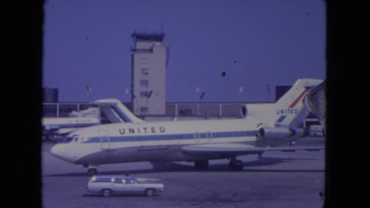 Side-on 1972 O'Hare view of a United jet parked near the control tower, with a small ground vehicle crossing the foreground.