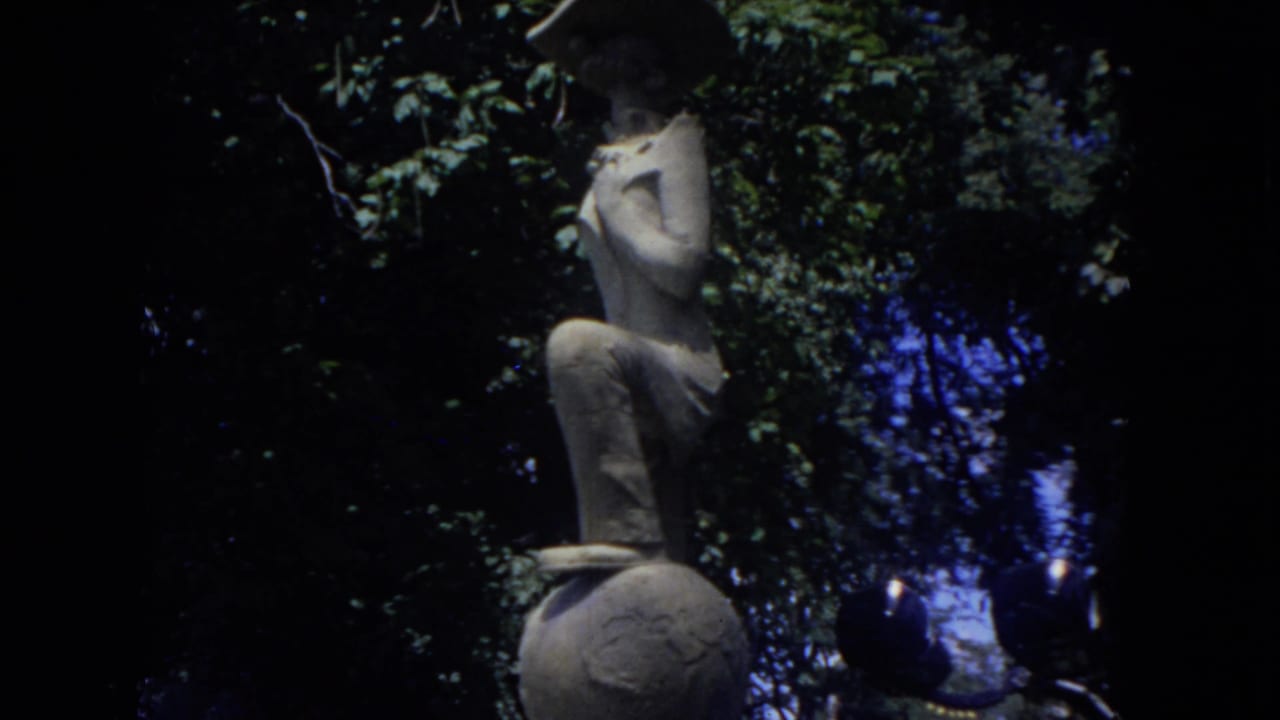 Cutaway from the airport footage to a stone statue of a figure poised on a ball, framed by leafy trees in the same 8mm reel.