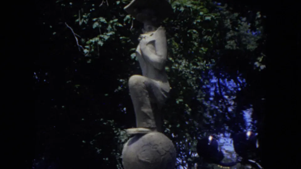 Cutaway from the airport footage to a stone statue of a figure poised on a ball, framed by leafy trees in the same 8mm reel.