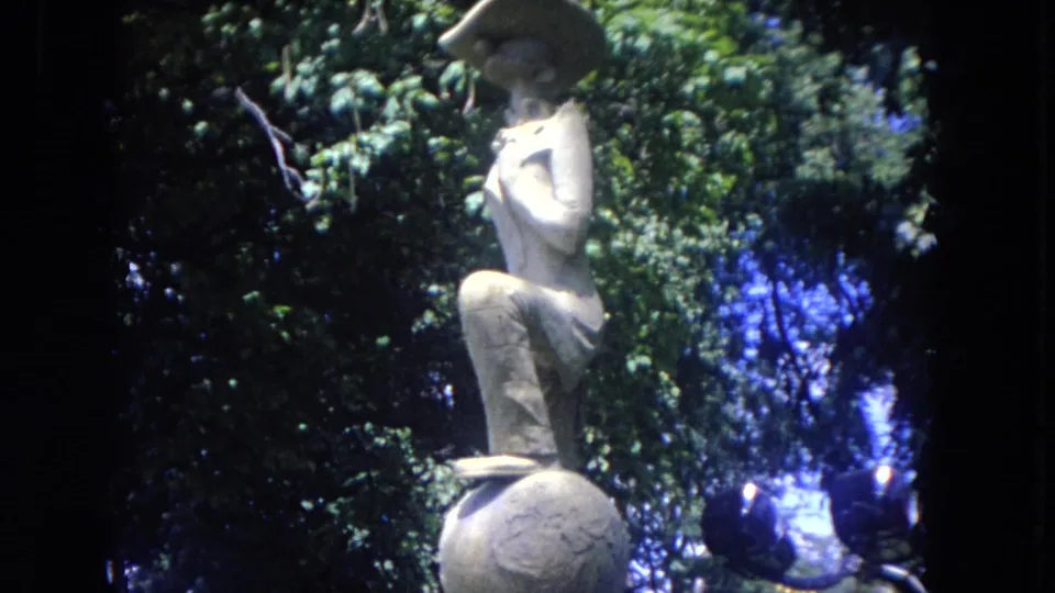 1972 8mm frame of a stone sculpture among dense trees near the Mississippi River waterfront.